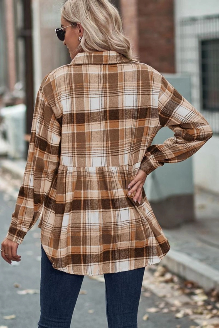 A woman with blonde hair, wearing a brown and white plaid long-sleeved shirt with a peplum hem and blue jeans, stands with her back to the viewer on a paved surface outdoors. She is also wearing sunglasses.