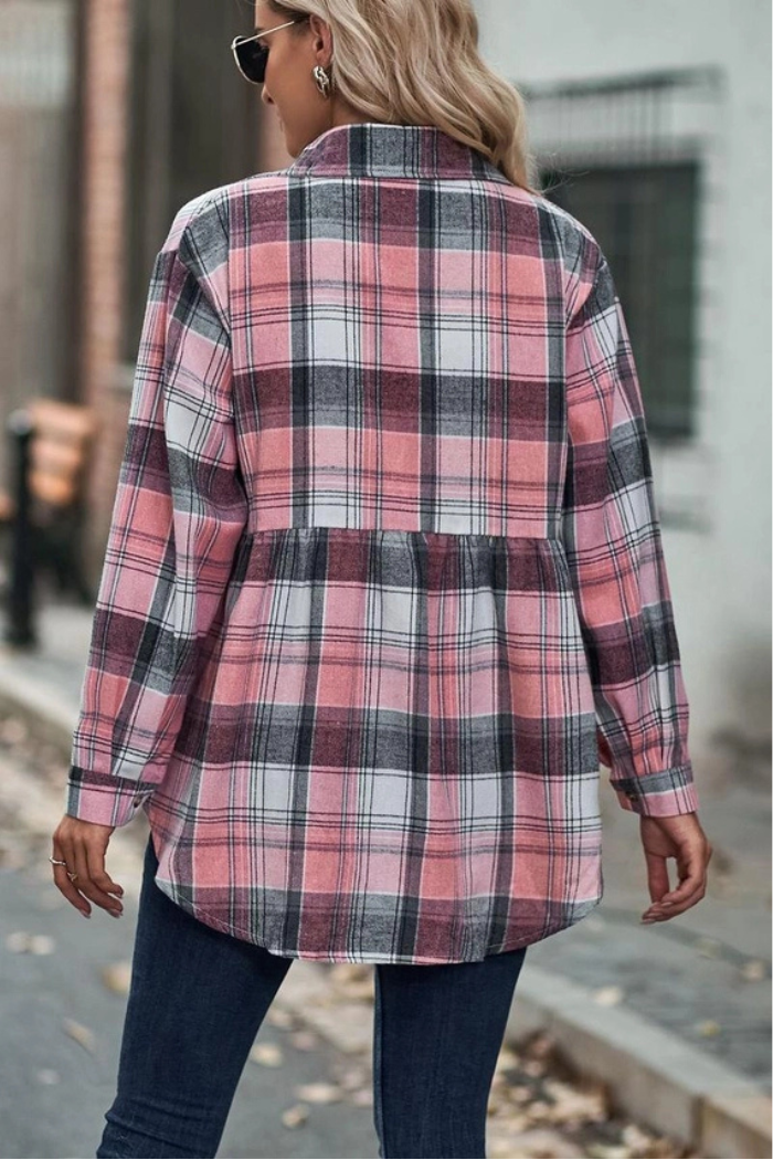 A woman viewed from the back, wearing a long-sleeved, pink, black, and white plaid shirt with a gathered peplum hem, paired with dark blue jeans. Her light-colored hair flows down her back, and she is outdoors on what appears to be a paved path with blurred buildings in the background.