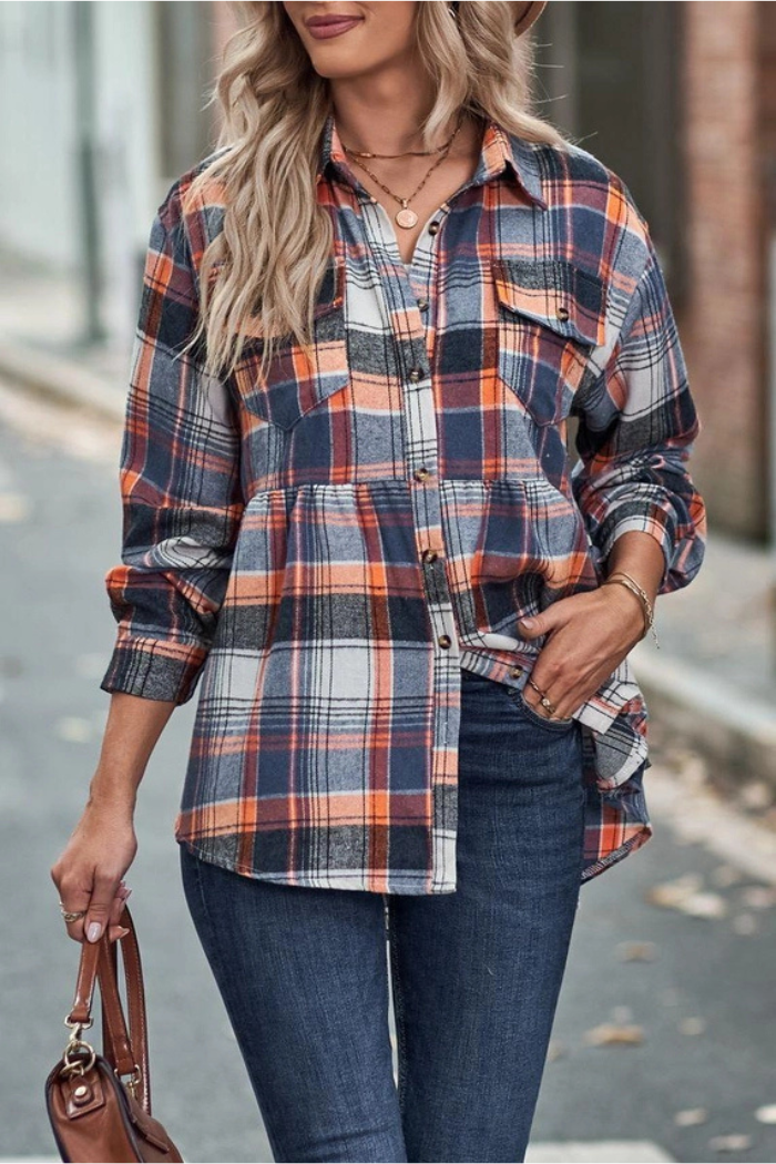 A woman with blonde hair wearing a blue, orange, and white plaid flannel shirt and dark blue jeans, holding a brown purse, stands on a street with blurred background foliage.