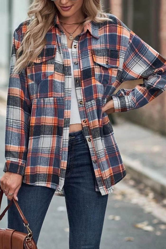 A woman with long blonde hair, wearing a long-sleeved blue, orange, and white plaid flannel shirt over a white top, paired with blue jeans. She is holding a brown bag in her left hand and posing outdoors.