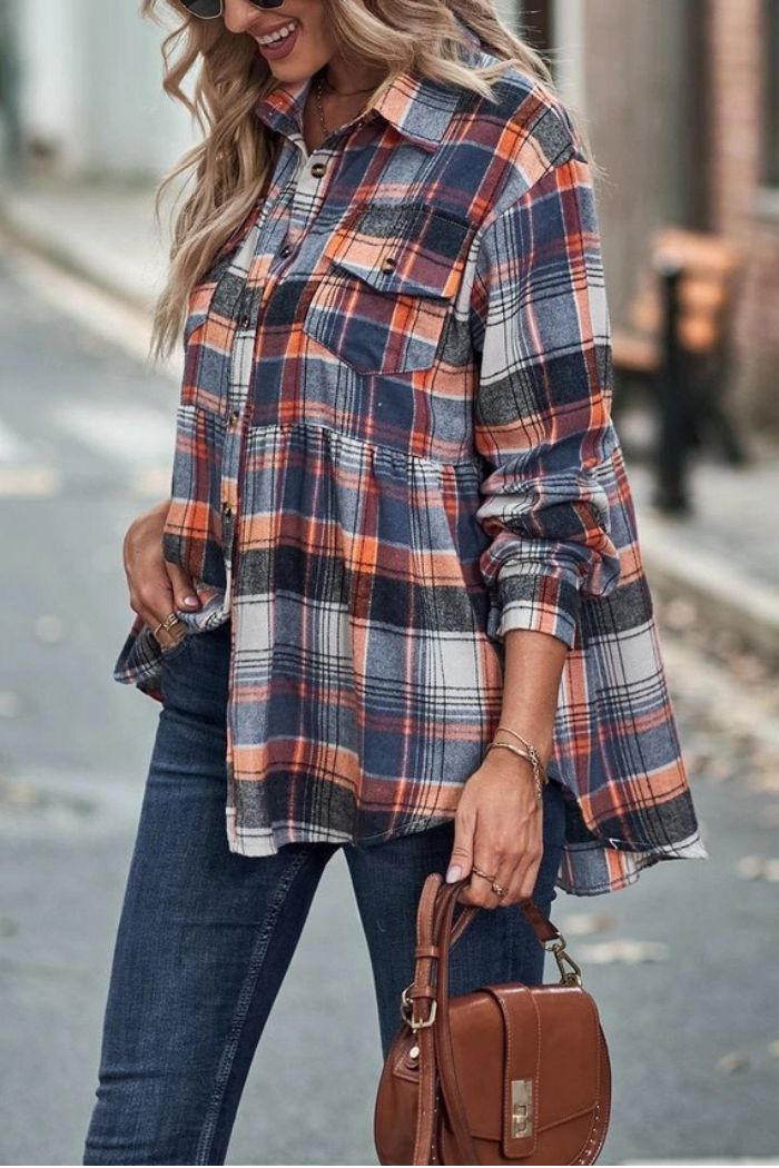 A woman walks outdoors wearing a long-sleeved, collared plaid shirt in shades of orange, navy, and white with a peplum hem, dark blue skinny jeans, and carrying a brown handbag. She has long, wavy blonde hair and sunglasses on her head.