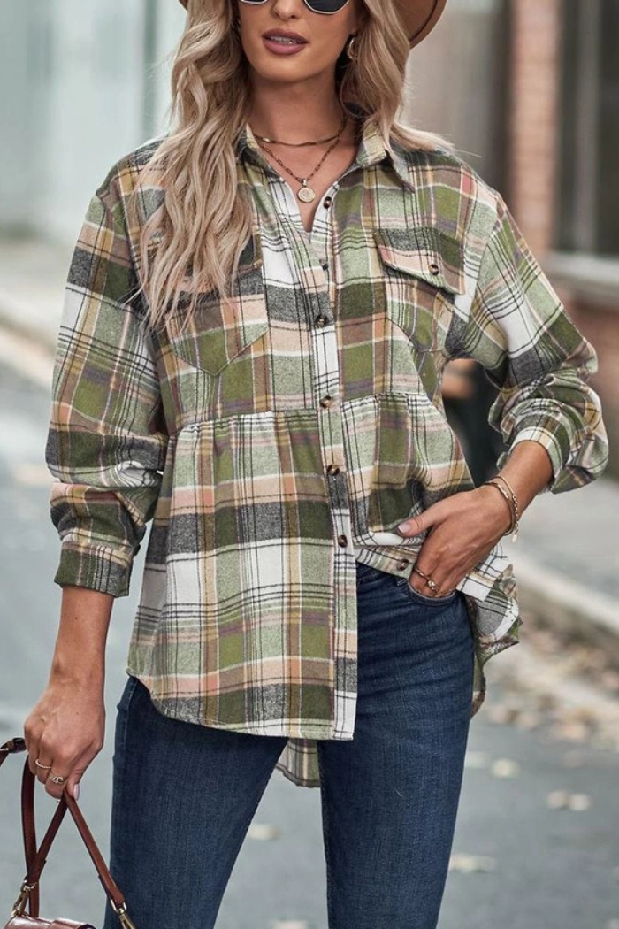 A woman with long blonde hair and a hat wears a casual green, white, and brown plaid button-down shirt with a peplum hem and long sleeves, paired with dark blue jeans, while holding a handbag and standing outdoors.