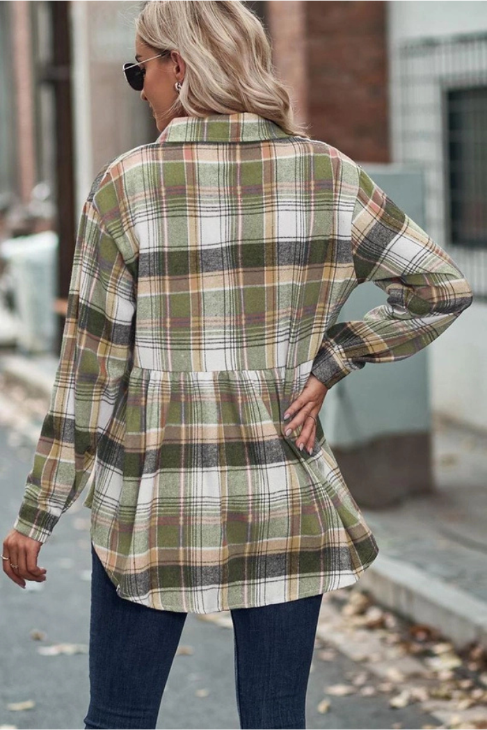 A woman from the back, wearing a green, white, and brown plaid flannel shirt with a peplum hem and long sleeves, paired with dark blue jeans. She is wearing sunglasses, and her blonde hair is down. The background is a blurred outdoor setting.