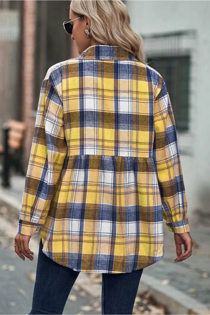 A person is shown from the back wearing a long-sleeved, plaid shirt in shades of yellow, blue, and white. The shirt has a gathered or peplum-style seam across the back at waist level. The person is standing outdoors, possibly on a street or sidewalk, and is also wearing dark pants and sunglasses on their head.
