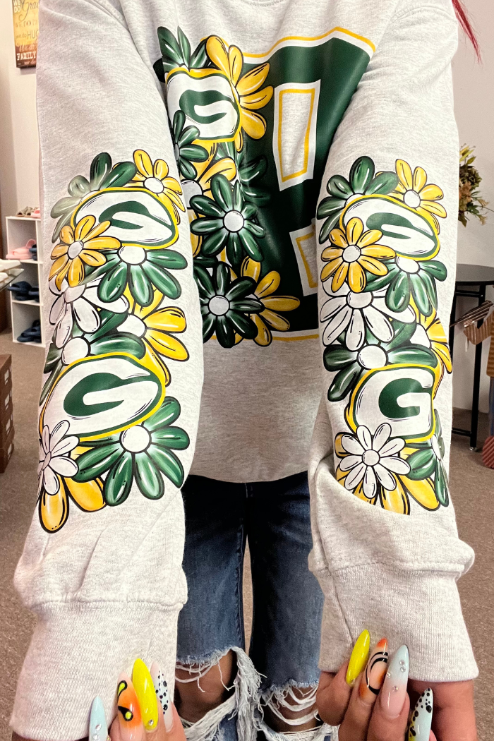 A person wearing a gray crewneck sweatshirt featuring a large green and yellow "G" logo, likely representing the Green Bay Packers, on the chest. The sleeves of the sweatshirt are adorned with a floral pattern composed of green leaves and yellow and white daisy-like flowers, interspersed with smaller "G" logos. 