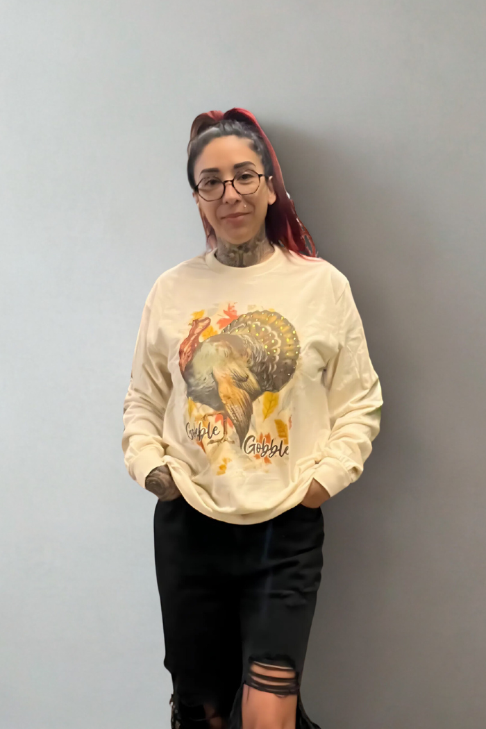 A woman with red hair and glasses, wearing a light-colored sweatshirt featuring a large turkey graphic and the words "Gobble Gobble," stands with her hands in the pockets of her ripped black jeans. She has visible tattoos on her arms and neck.
