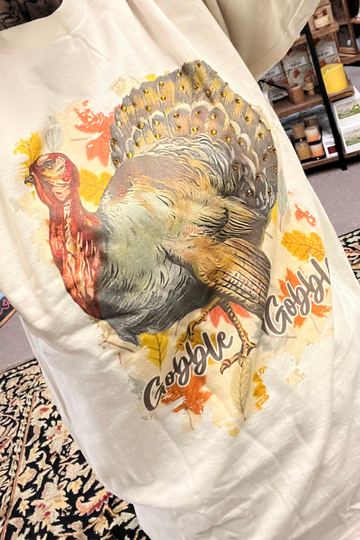 A beige t-shirt featuring a graphic of a turkey with gold embellishments on its feathers, surrounded by colorful fall leaves in shades of orange, yellow, and brown. The text "Gobble Gobble" is written in a stylized font below the turkey. 