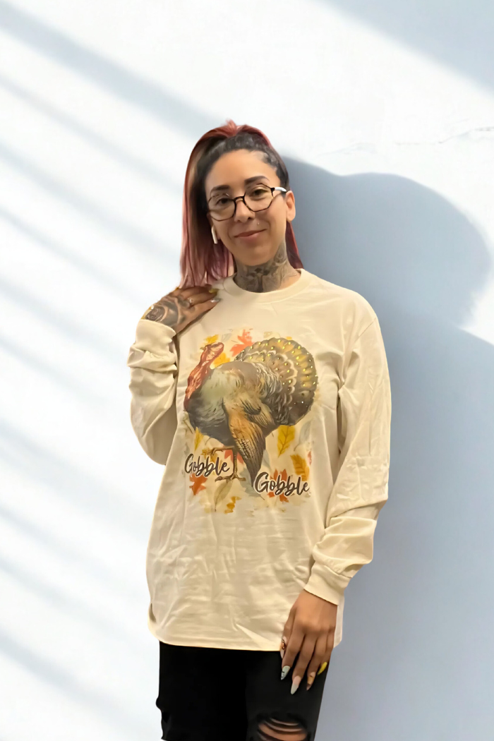 A woman with dark hair and glasses, wearing a light-colored long-sleeved t-shirt with a large graphic of a turkey surrounded by autumn leaves and the words "Gobble Gobble" printed on it. The woman is smiling slightly and has visible tattoos on her neck and arm.