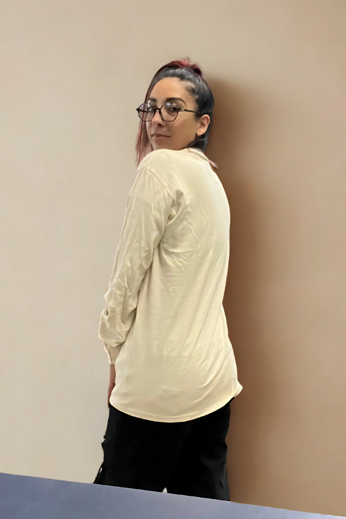 A young woman with dark hair pulled up and subtle red highlights wears glasses, a cream-colored long-sleeved shirt, and black pants, looking back over her left shoulder at the camera in front of a neutral-toned wall.