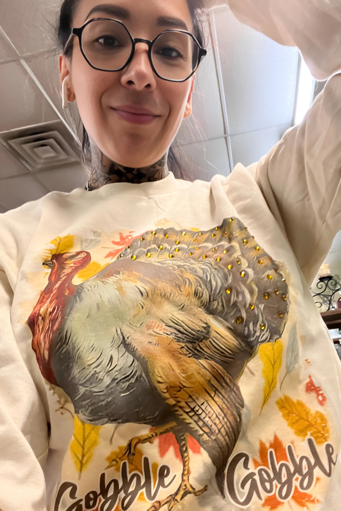 A close-up selfie shows a person with dark hair, glasses, and a visible tattoo on their neck, smiling at the camera. They are wearing a light-colored sweatshirt featuring a large, colorful graphic of a turkey adorned with small golden embellishments. The design also includes autumn leaves and the phrase "Gobble Gobble" printed below the turkey.
