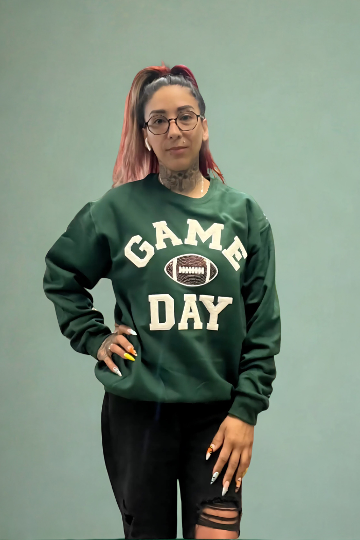 A woman with dark hair tied in a ponytail, wearing glasses and a dark green crewneck sweatshirt with "GAME DAY" in white varsity-style letters and a football graphic in the center, stands with her left hand on her hip against a light blue-green wall. 