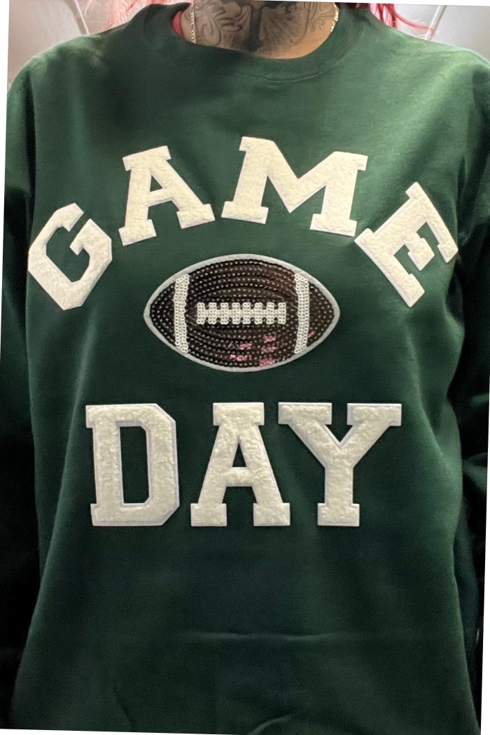 A person wearing a dark green sweatshirt with "GAME DAY" in white letters and a sequined football graphic in the center.
