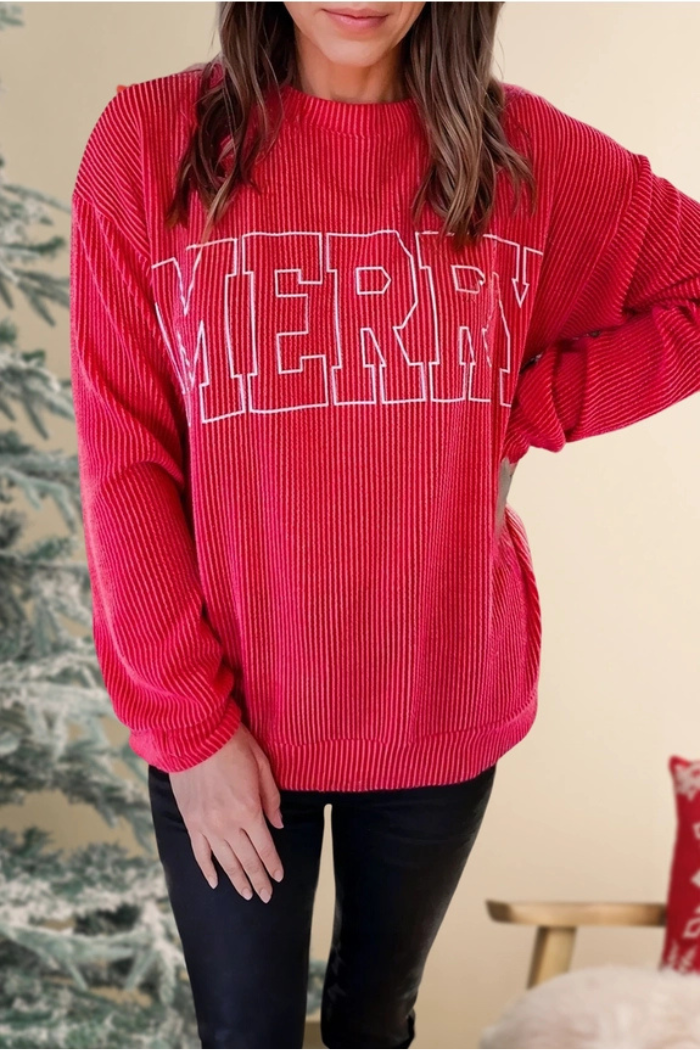 A person wearing a red, ribbed, long-sleeved crewneck sweater with "MERRY" outlined in white on the front, paired with black pants. A blurred Christmas tree is visible in the background.
