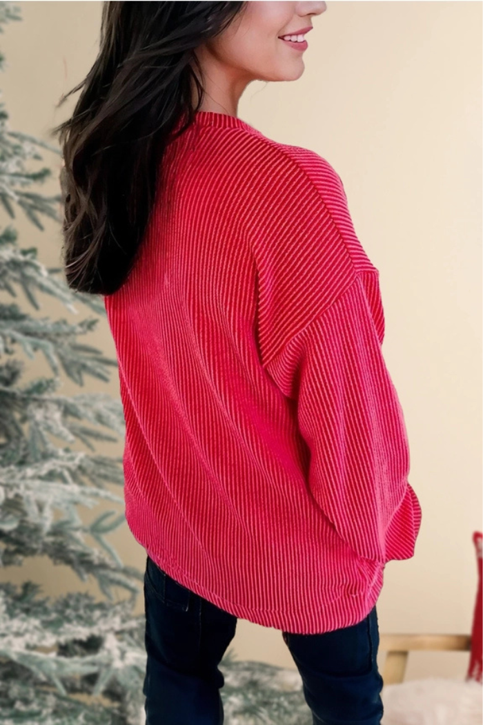 A woman from the back wearing a red ribbed long-sleeved top, slightly turned to show her profile, with a blurred background featuring what appears to be a decorated Christmas tree. 