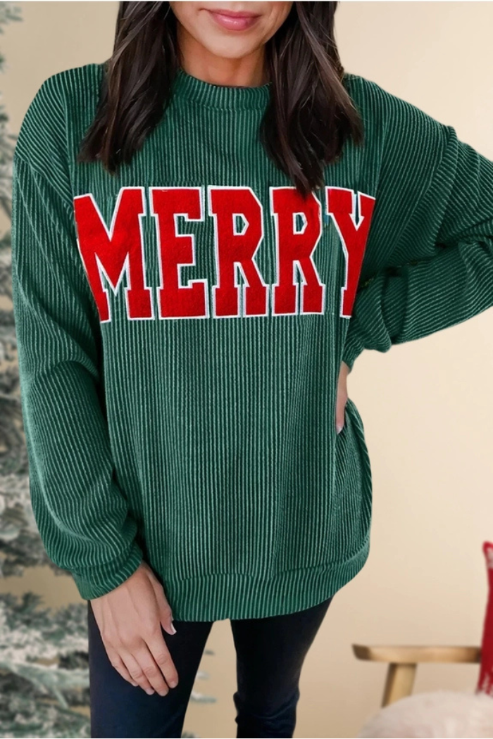 A woman models a long-sleeved, loose-fitting, dark green ribbed top with the word "MERRY" emblazoned in large, bold red letters across the chest.