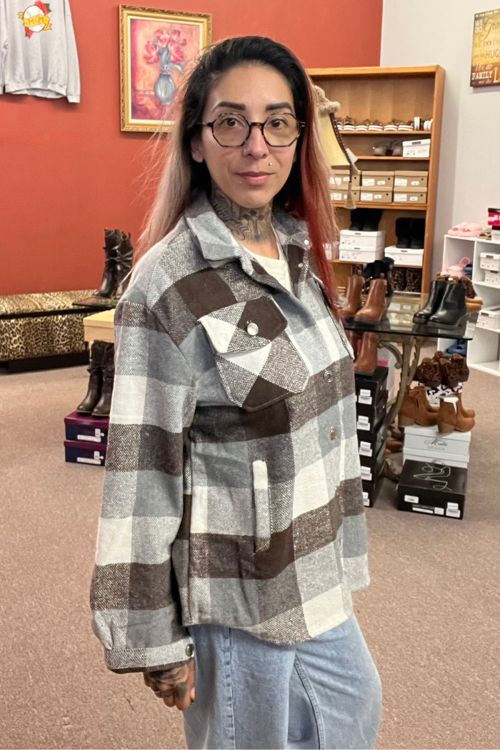 A woman with glasses and tattoos on her neck and hands stands in a store, wearing a grey, white, and brown plaid shacket over a white shirt and light blue jeans. She is looking directly at the camera. In the background, there are shelves of boots and a framed painting on the wall.
