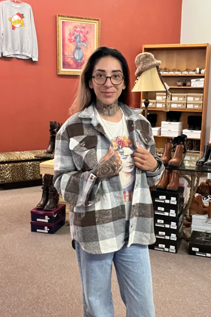 A woman with glasses and tattoos stands in a store, wearing a gray, white and brown plaid shacket over a white t-shirt with a colorful graphic print. She is holding the front of the shacket open with her hands.