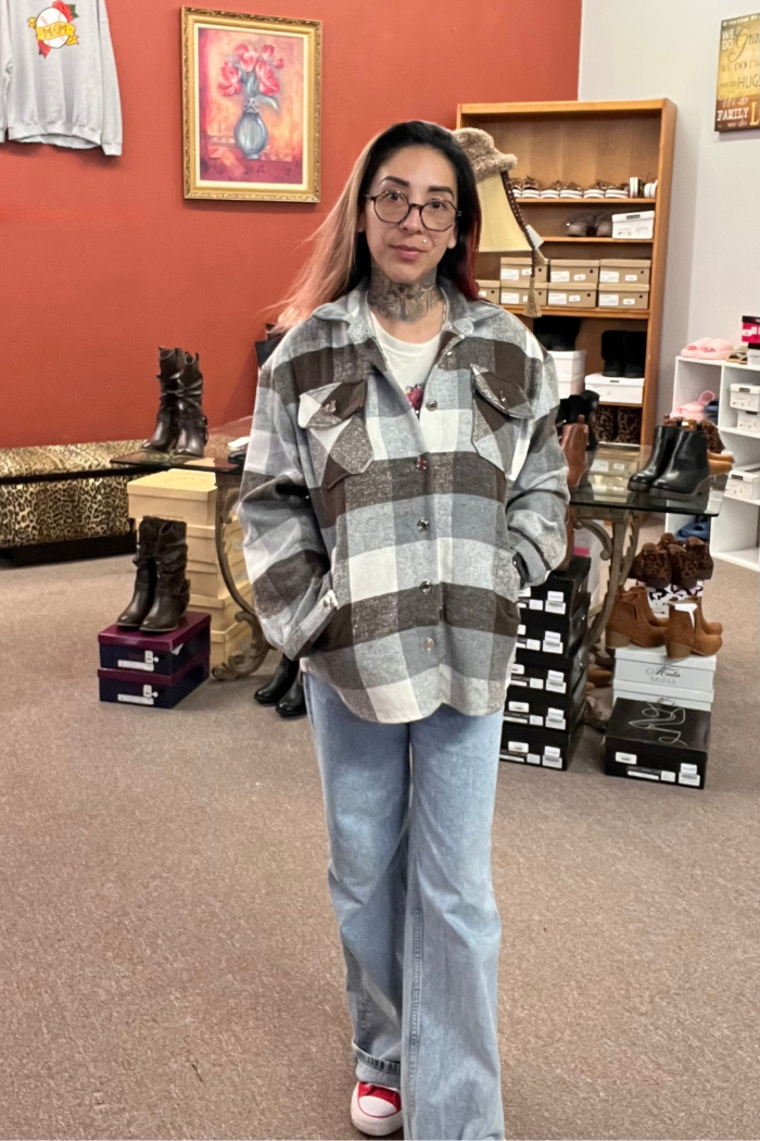 A woman with long dark hair and glasses stands indoors, wearing a grey, white and brown plaid shacket over a white t-shirt and light blue jeans. She has a tattoo on her neck, and her hands are in the pockets of the shacket.