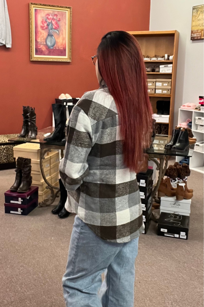 A person with long reddish-brown hair is shown from the back, wearing a gray, white, and brown plaid flannel shacket over blue jeans. The person is standing inside what appears to be a clothing store, with shoes and other merchandise visible in the background.