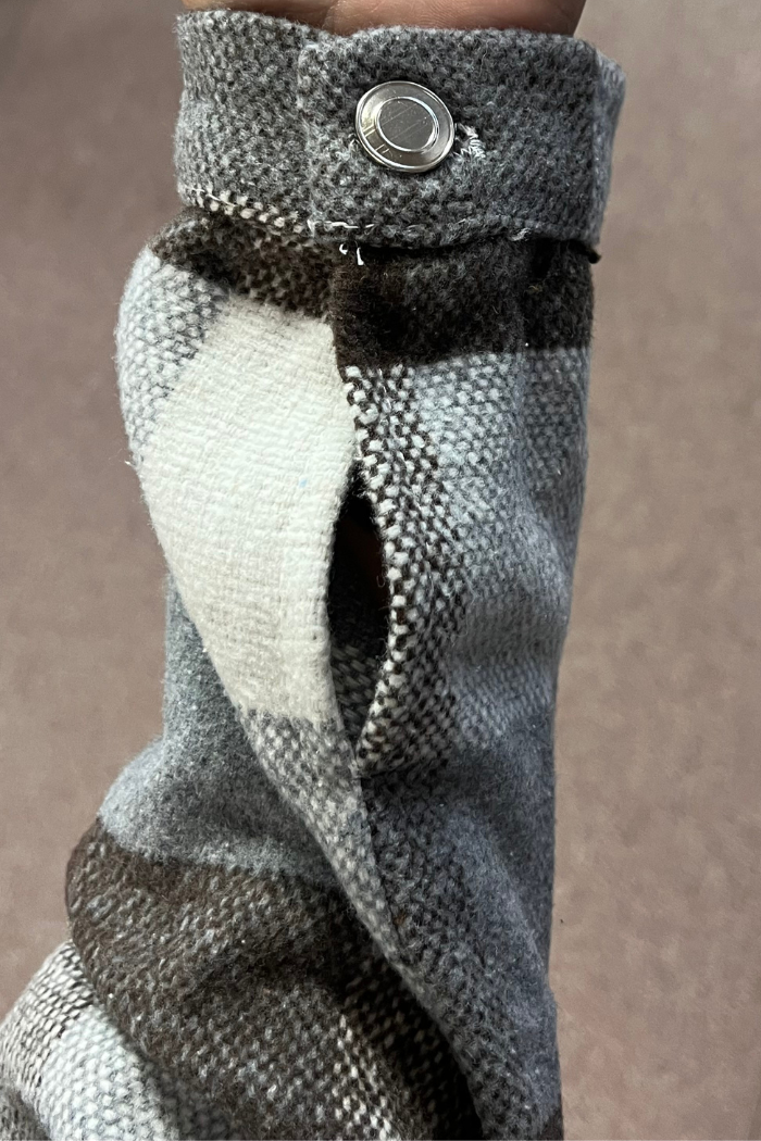 A close-up shot of a person's arm wearing a gray, white, and brown plaid shacket. The sleeve is held in place by a button tab with a silver snap button.