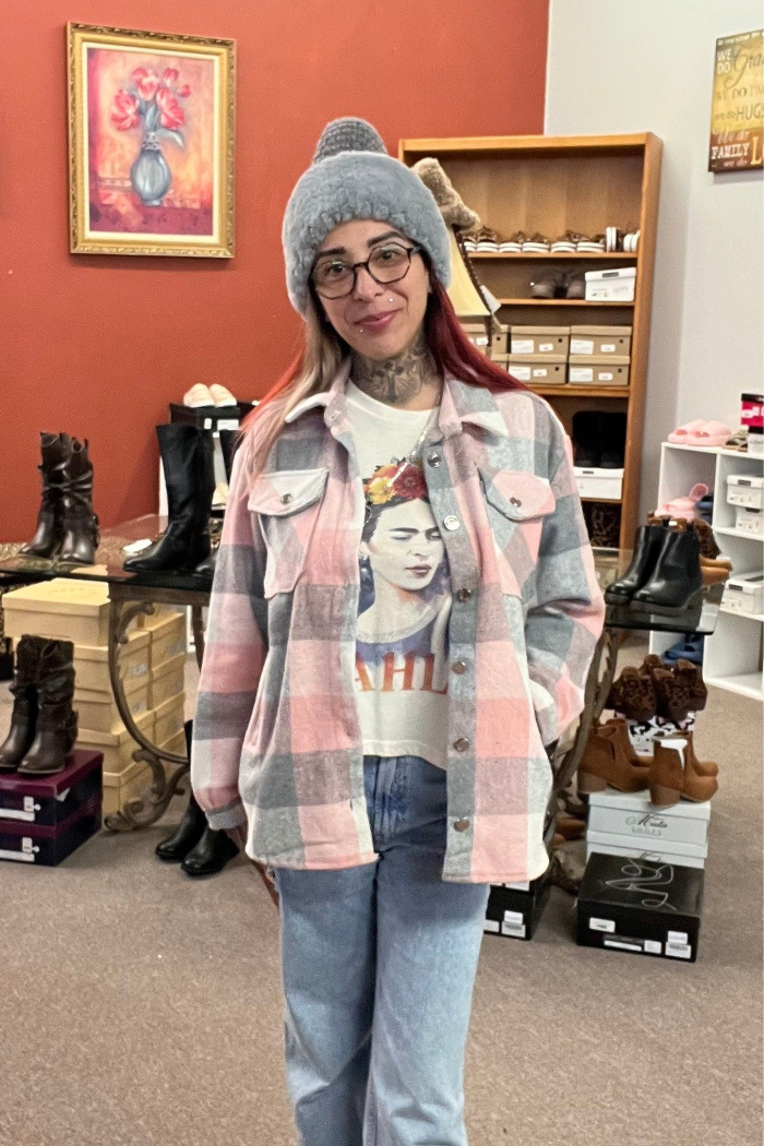 A woman is wearing a grey beanie, glasses, a light-colored t-shirt with a graphic of Frida Kahlo, and a pink, white and grey plaid flannel shacket over it. She is also wearing blue jeans. In the background, there are shelves with shoes and a painting on the wall.