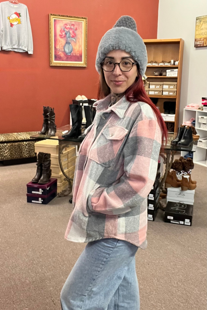 A woman wearing a gray beanie and a pink, white and gray plaid flannel shacket stands with her hands in her pockets, looking at the camera. She is indoors, and there are boots and shelves with boxes in the background.