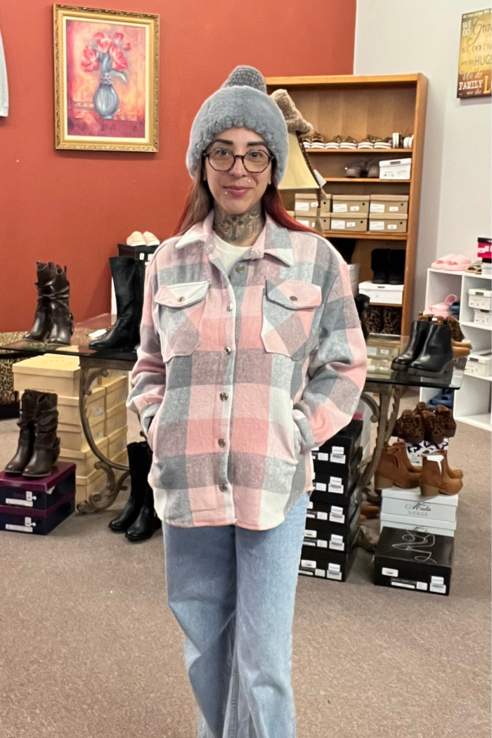 A woman is wearing a pink, grey, and white plaid shacket over a white t-shirt and light-wash blue jeans. She is wearing glasses and a grey beanie.
