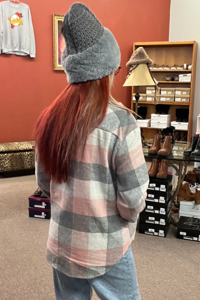 A woman shown from behind, wearing a gray, white and pink plaid flannel-style shacket. She is also wearing a gray knit beanie and light-wash blue jeans. The person is standing in what appears to be a clothing boutique, with various shoes and other items visible in the background.