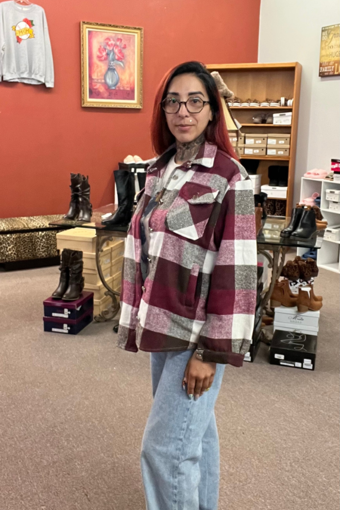 A woman with reddish-brown hair and glasses stands in a store, wearing a burgundy, white, and brown plaid flannel-style shacket, a white shirt, and light-wash blue jeans. She has a tattoo on her neck and is standing in front of shelves of shoes and other merchandise.
