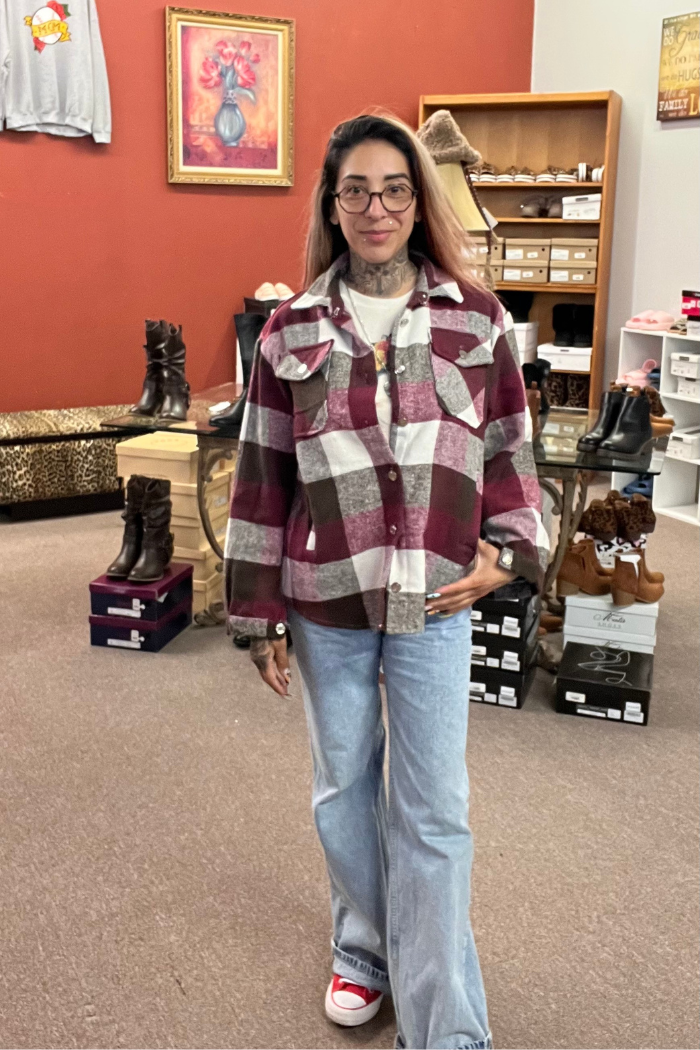 A woman with long dark hair and glasses is standing indoors, wearing a burgundy, white, and gray plaid flannel shacket, a white t-shirt, light blue wide-leg jeans, and red Converse sneakers. She has her left thumb in pocket. The background appears to be a store with shoes and other items on shelves.