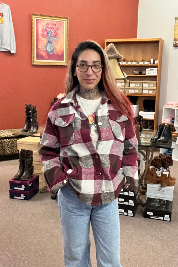 A woman wearing a burgundy, white, and brown plaid flannel shacket, a white t-shirt, and light blue jeans. She is looking directly at the camera with a slight smile. Both hands in her jean pockets. The background shows a painting on the wall and various footwear.