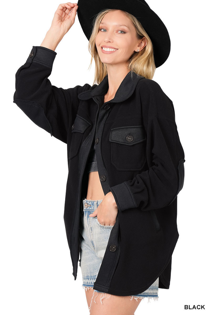 A smiling blonde woman wearing a black hat and a black, oversized fleece shacket with button closures and elbow patches. She is also wearing light-wash denim shorts. The text "BLACK" is visible in the bottom right corner of the image. 