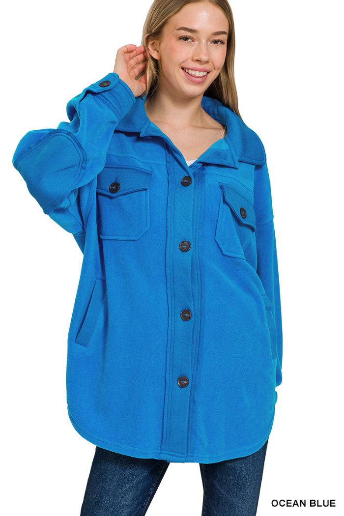 A blonde woman with a bright smile models a blue fleece shacket. The jacket is oversized, has two large chest pockets, and two side pockets. The sleeves have elbow patches, and the front has several dark brown buttons.
