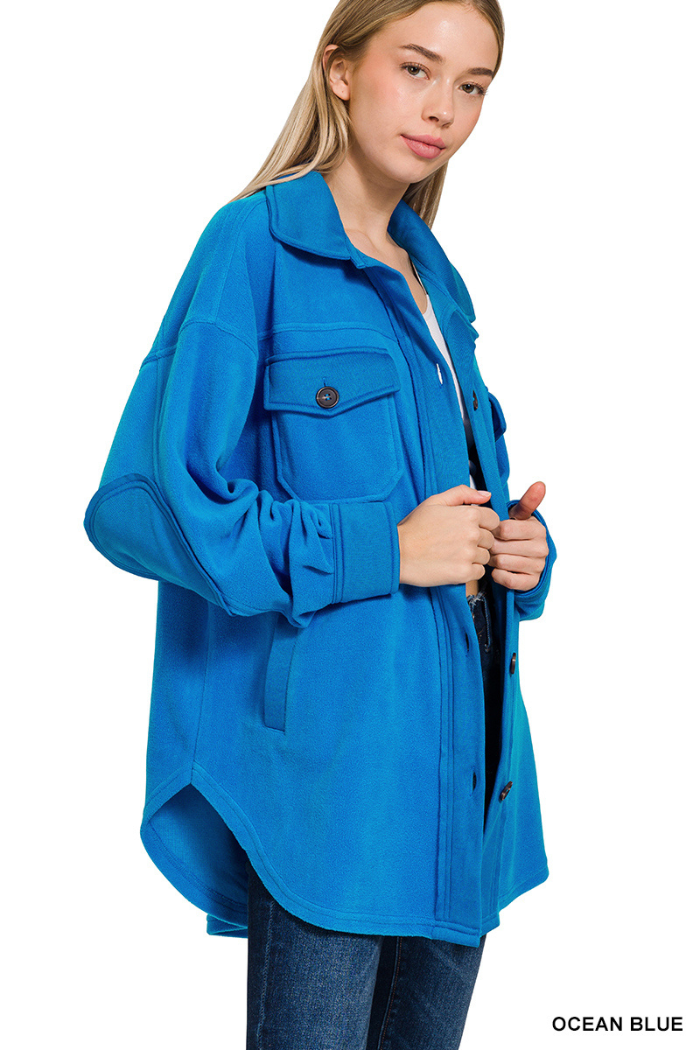 A woman wearing a bright blue fleece shacket with an oversized fit. The shacket has a collar, two large chest pockets with buttons, and long sleeves with elbow patches. The woman is holding the front of the shacket with her hands.