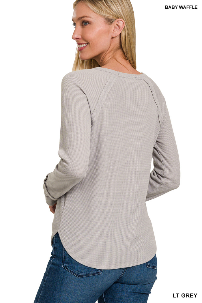 A woman with long blonde hair models the Waffle Knit Long Sleeve Top in light grey, featuring a rounded hem, long sleeves, and a casual fit, shown from the back and slightly angled, paired with classic blue jeans.