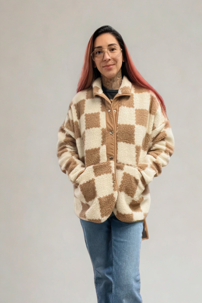A woman with long hair and glasses is smiling at the camera. She is wearing a brown and white checkered sherpa jacket and blue jeans, with her hands in the jacket's pockets.