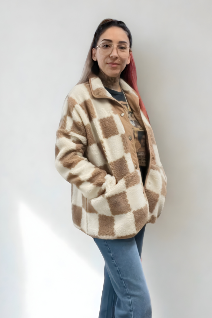 A woman with glasses and tattoos is standing against a white wall, wearing a checkered brown and white sherpa jacket. She has her hands in the pockets of the jacket and is looking at the camera.