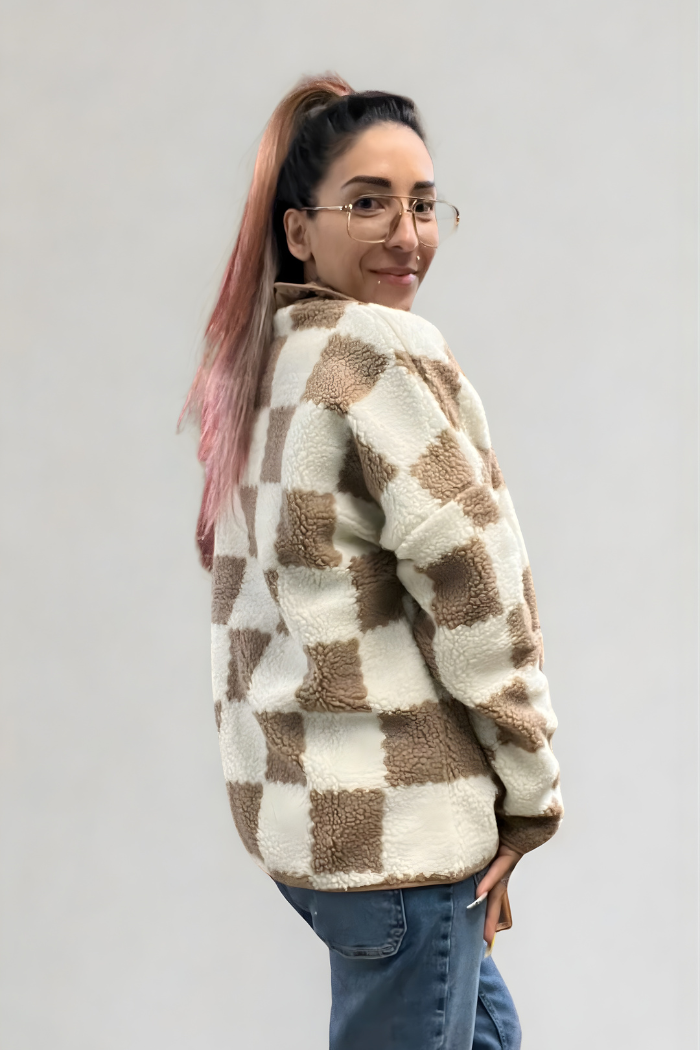 A woman with glasses and pink-streaked hair in a ponytail smiles while looking over her shoulder. She is wearing a checkered brown and white sherpa jacket over a dark shirt and blue jeans.