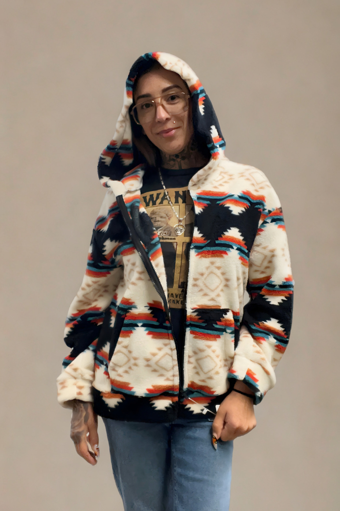 A woman with tattoos on her neck and arms is wearing a white, black, and orange sherpa hoodie with a southwestern or Aztec pattern. She is wearing glasses and a necklace over a dark t-shirt. The hoodie has a full zipper and a hood with a similar pattern.