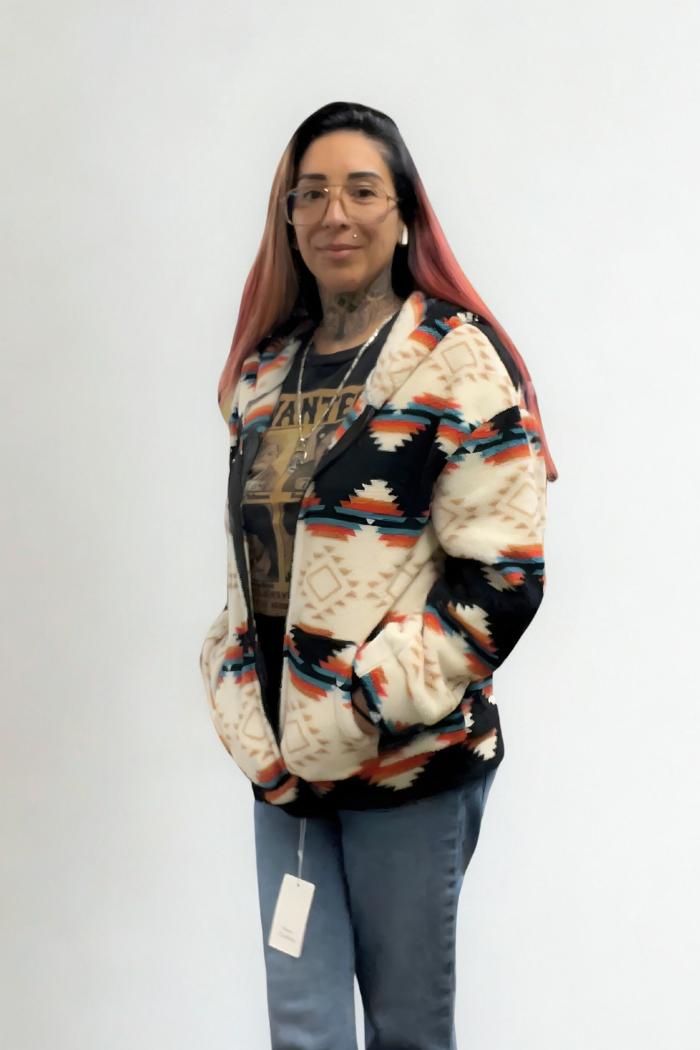A woman with long dark hair with pinkish ends and glasses stands against a white background, wearing a black t-shirt with a graphic design, a cream-colored sherpa shacket with a black, red, and turquoise Aztec pattern, and blue jeans.