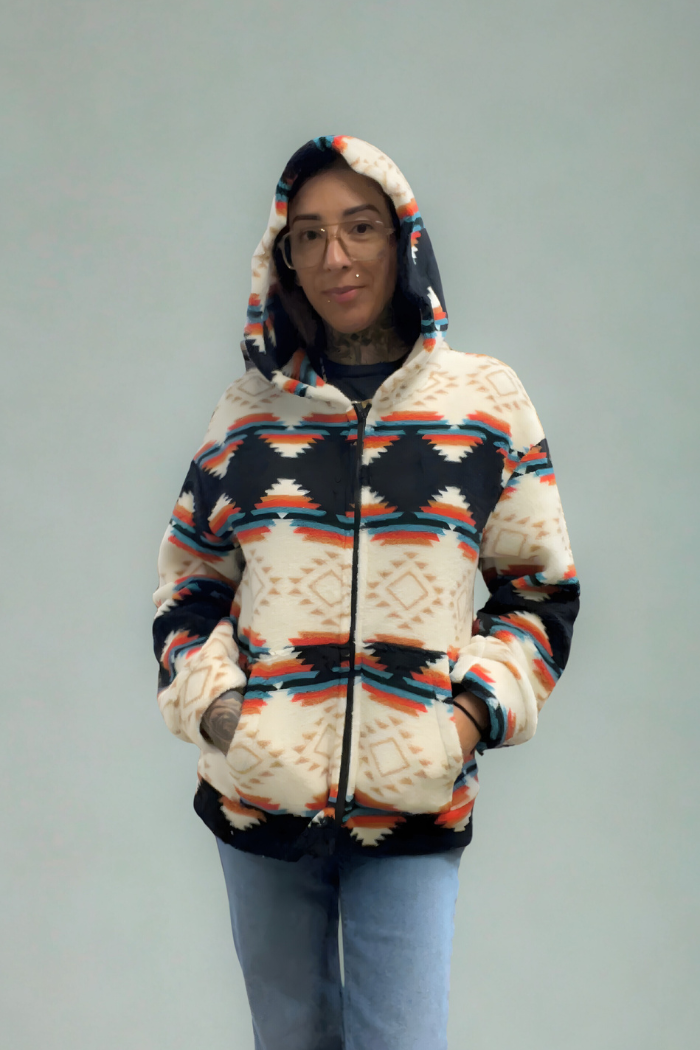 A person with glasses and tattoos on their arms is wearing a hooded fleece shacket with a distinctive geometric pattern in white, navy blue, orange, and light blue, and blue jeans. The person is standing against a plain, light-colored wall with their hands in the shacket pockets.