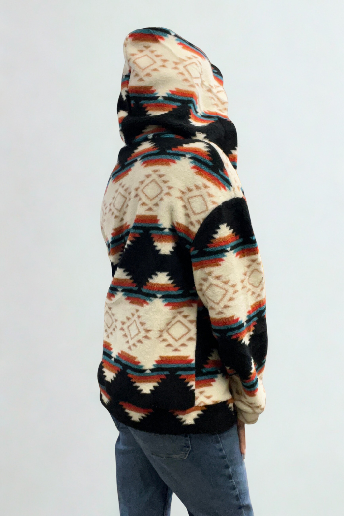 A person wearing a hooded fleece shacket with a black, cream, orange, and turquoise geometric pattern. The person is shown from the back, with their hood up, and is also wearing blue jeans. The shacket has a zip-up front and pockets.