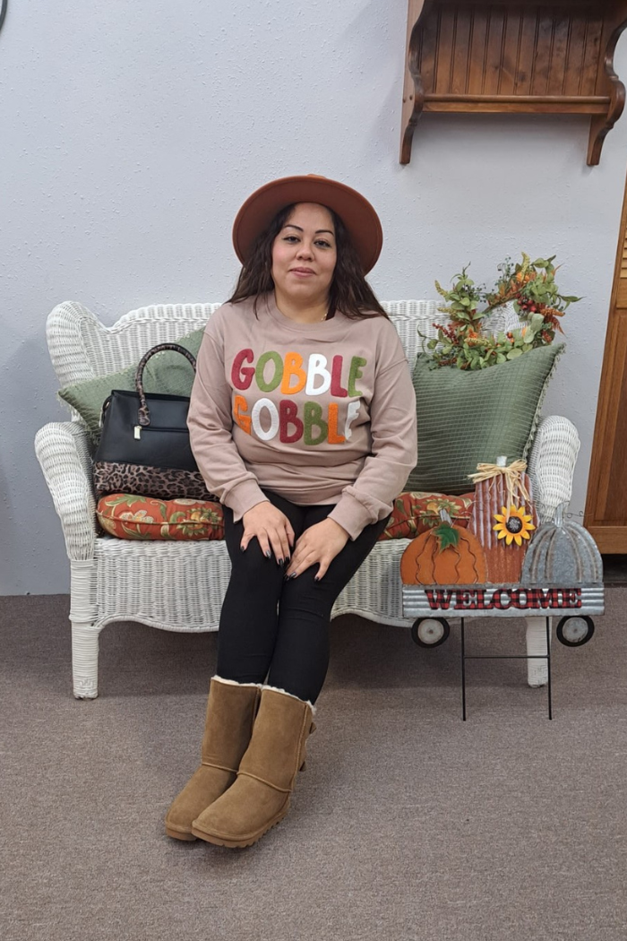 A woman wearing a brown hat, a beige "Gobble Gobble" sweatshirt with colorful chenille letters, black leggings, and brown boots sits on a white wicker bench. A black handbag with a leopard-print strap is next to her. The bench is decorated with pillows, and a small cart with a pumpkin and a "WELCOME" sign is in front of it.
