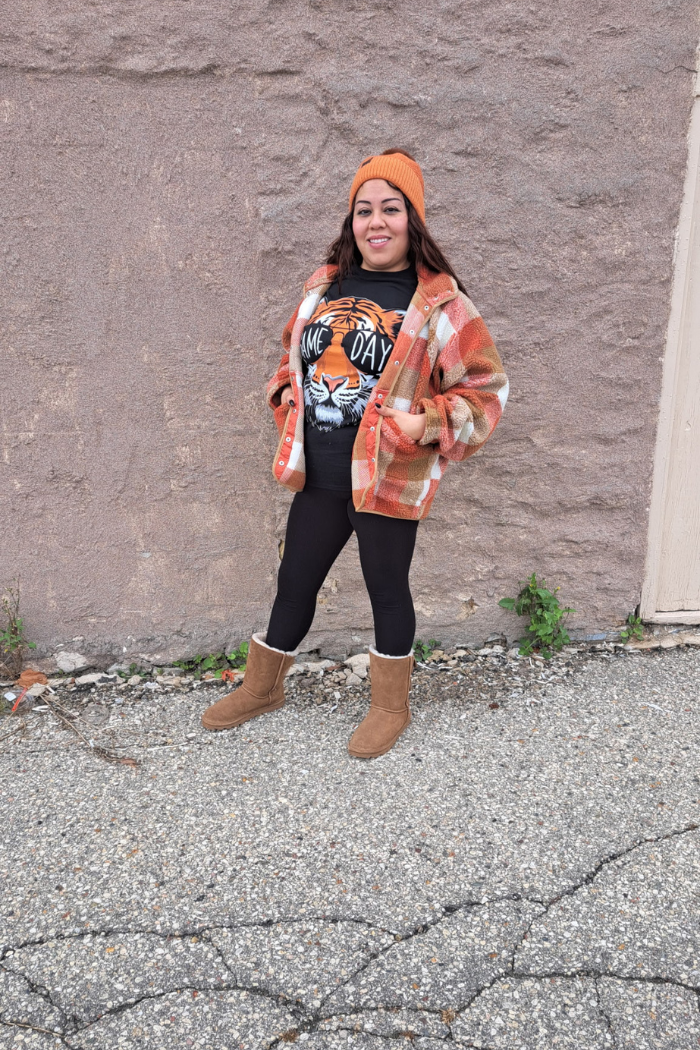 A woman stands in front of a stone wall, smiling at the camera. She is wearing a black t-shirt with a tiger head graphic and the words "GAME DAY" on it, an orange and white plaid flannel jacket, black leggings, and brown UGG-style boots. She also wears an orange beanie.