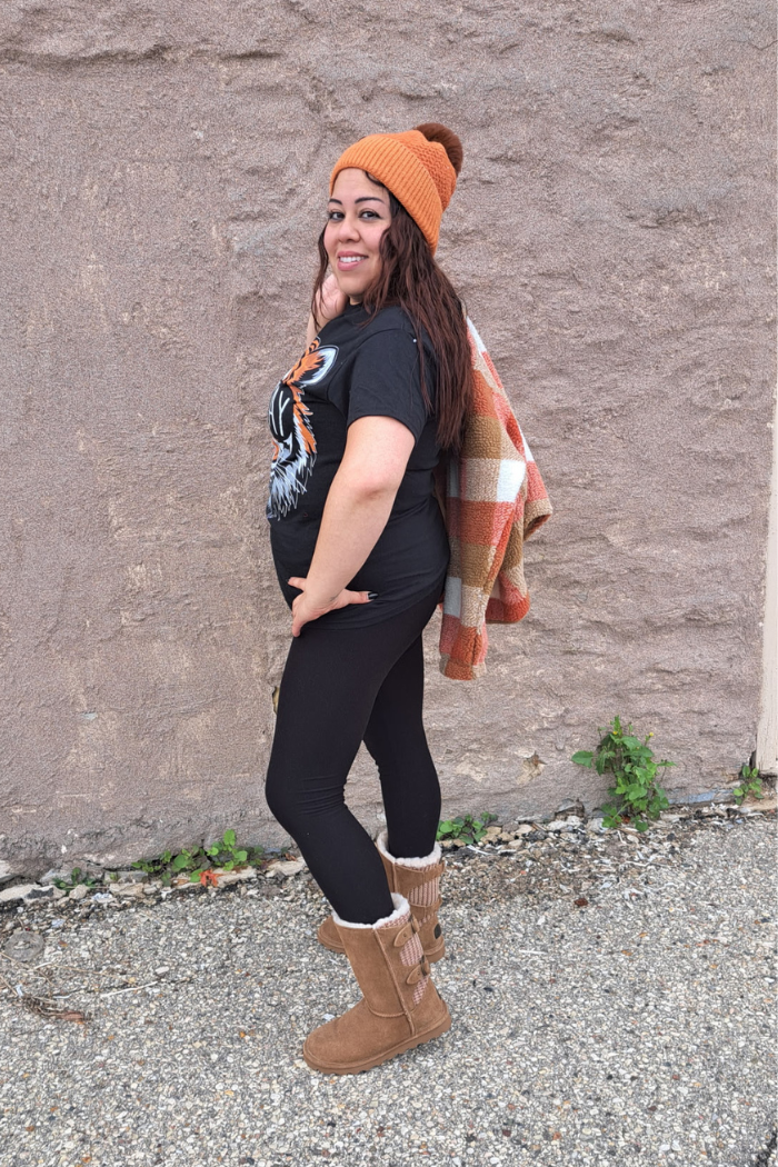 A woman stands sideways to the camera, smiling over her shoulder. She is wearing a black t-shirt with a tiger graphic, black leggings, and tan boots with faux fur trim. She holds a cream and orange plaid shacket over her shoulder and wears an orange beanie.