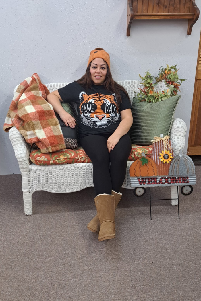 A woman with brown hair is seated on a white wicker bench. She is wearing a black t-shirt with a "GAME DAY" tiger graphic, black leggings, and tan UGG-style boots. A rust-and-tan plaid shacket is draped over the arm of the bench. The bench has orange and green cushions. To the right of the person, a decorative sign with "WELCOME" written on it features pumpkins and a sunflower. A fall-themed wreath and a striped pillow are also visible. The background is a plain gray wall and a wooden shelf.