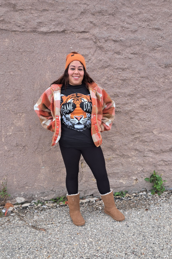 A smiling woman stands against a stone wall, wearing a black t-shirt with a tiger head graphic and the text "GAME DAY". She is also wearing a plaid flannel jacket, black leggings, an orange beanie, and tan boots.