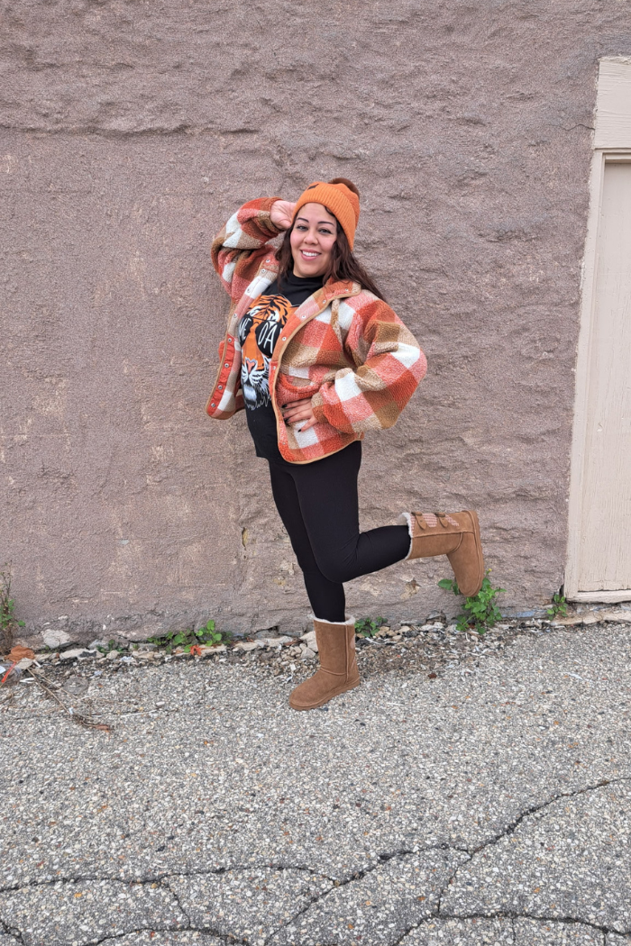 A woman wearing a plaid shacket, a black shirt with a tiger graphic, black leggings, and brown boots, and an orange beanie, poses in front of a stone wall.