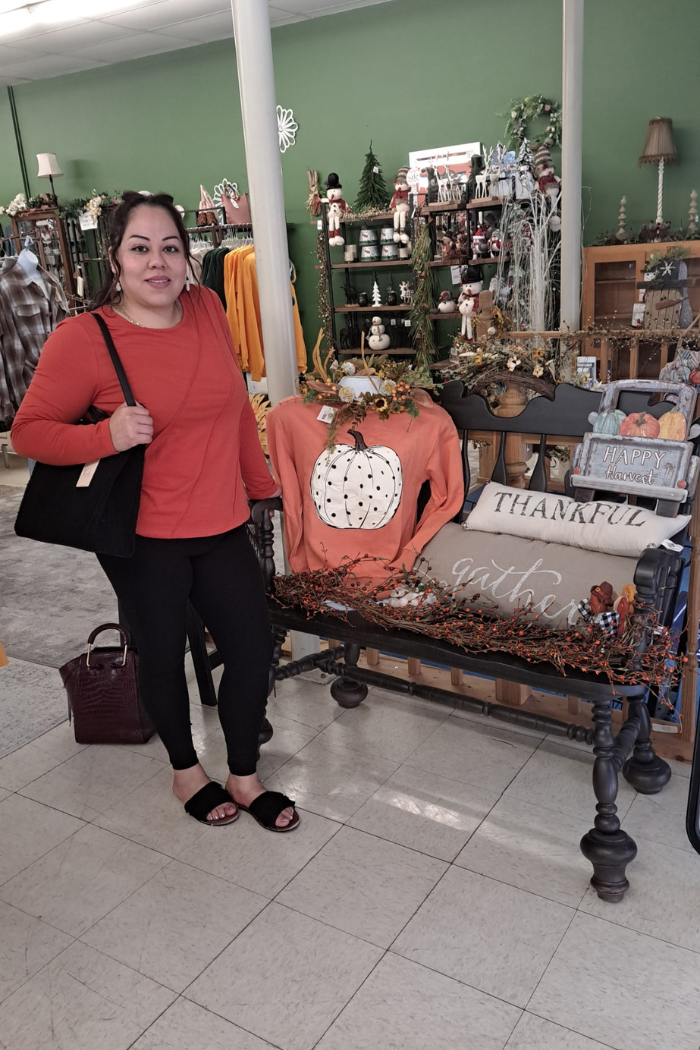 A woman in an orange shirt and black leggings stands inside a store next to a black bench. The bench is decorated with fall-themed items, including an orange sweater with a pumpkin design on a mannequin torso, pillows that say "THANKFUL" and "gather," and various garlands of berries and leaves. The store shelves in the background are filled with other seasonal decorations.