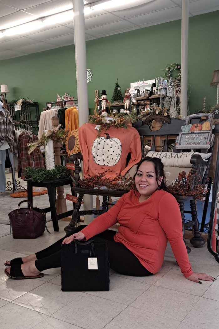 A woman with dark hair wearing a long-sleeved orange shirt and black pants is sitting on a tiled floor in a clothing store, smiling at the camera. In front of her is a black handbag. Behind her is a bench with autumn-themed decorations, including a pillow that says "THANKFUL" and a sweater with a pumpkin design on it. Clothing racks are visible in the background.